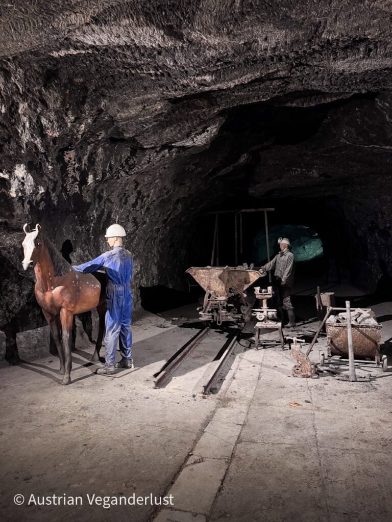 Eine Szene in einem historischen Bergwerk mit lebensgroßen Figuren von Bergleuten und einem Pferd, umgeben von Karren und Gleisen, dramatisch beleuchtet vor schroffen Felswänden.