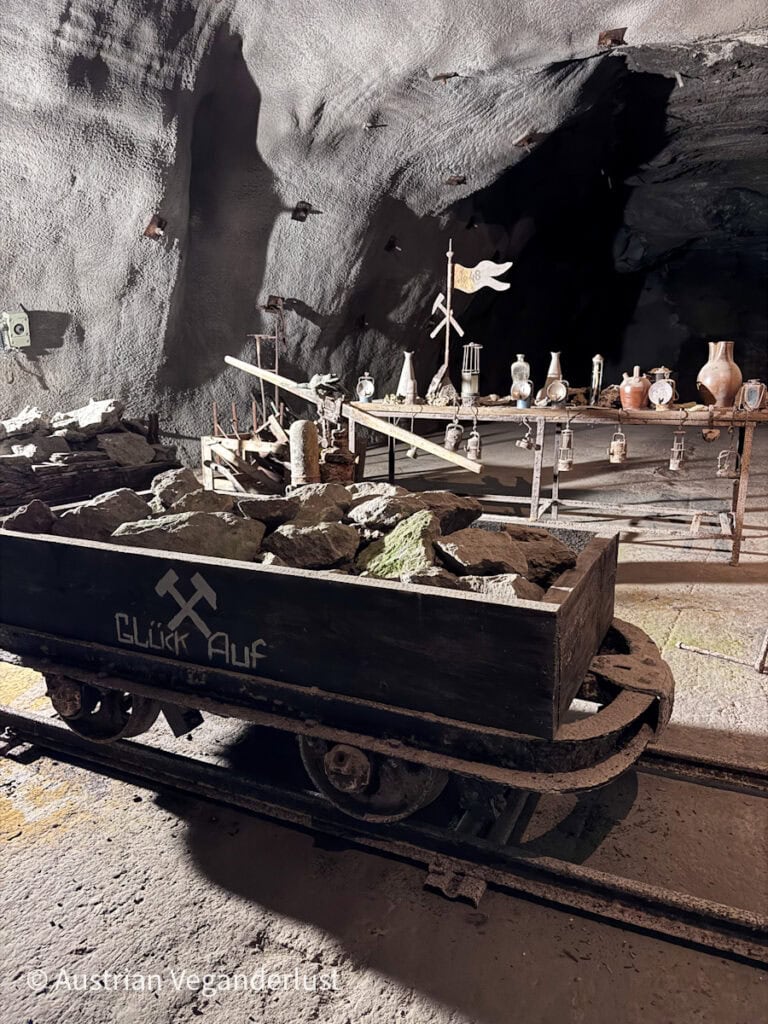 Ein mit Steinen beladener Grubenwagen mit der Aufschrift „Glück Auf“ vor einem langen Holztisch, auf dem alte Laternen, Werkzeuge und Töpferwaren ausgestellt sind, in einem schummrigen Tunnel.