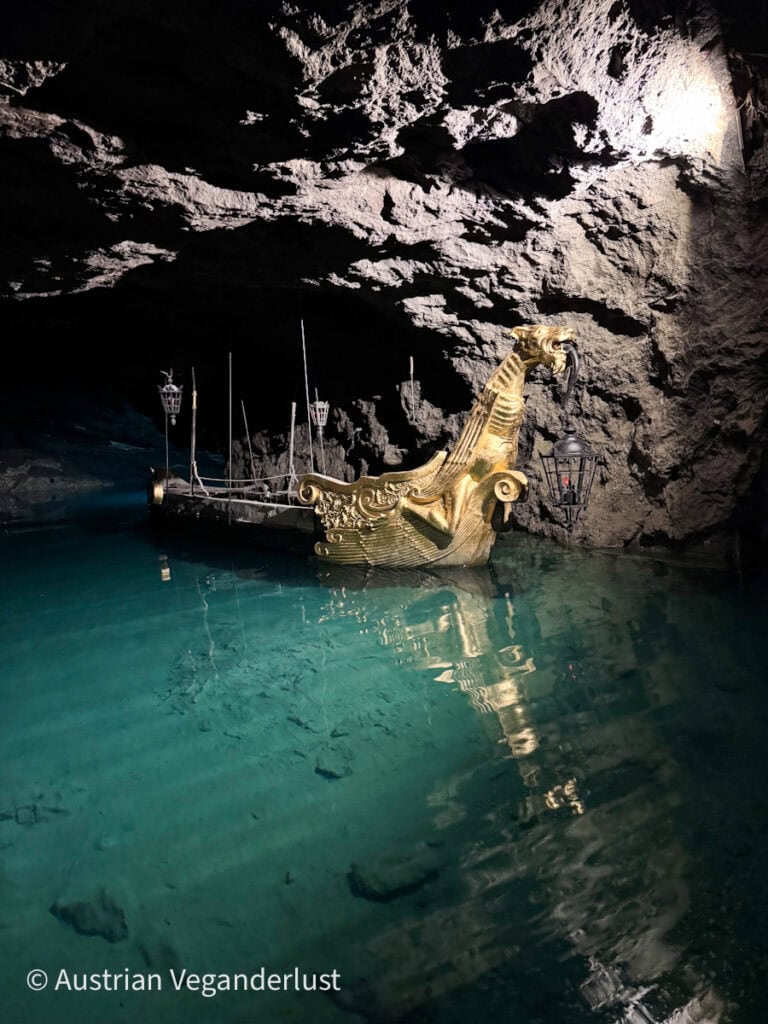 Ein goldenes, drachenförmiges Zierboot schwimmt auf einem klaren, blauen unterirdischen See in einer felsigen Höhle, umgeben von Laternen.
