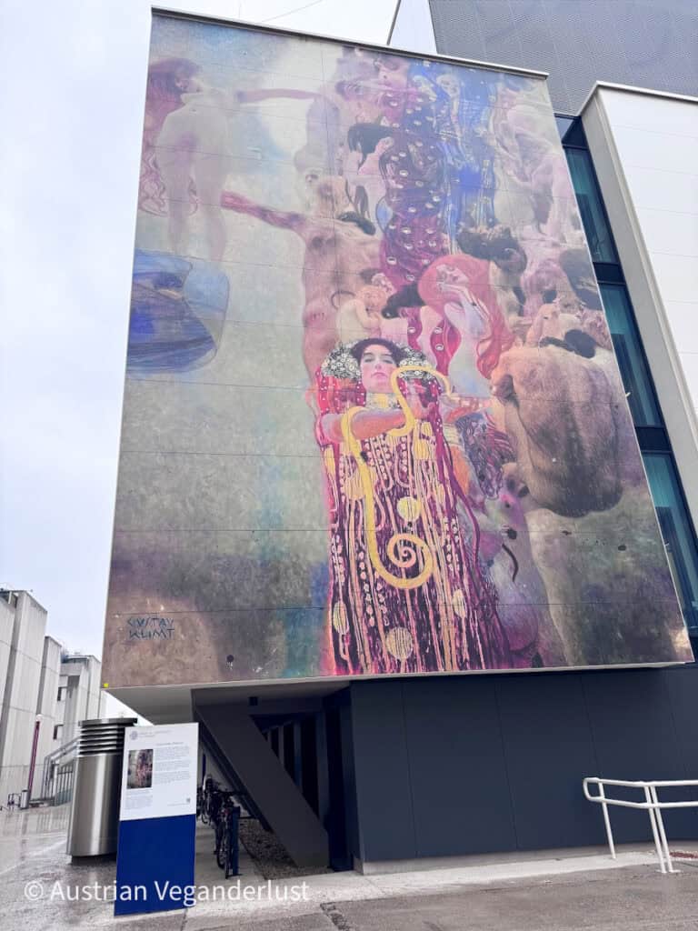 Eine großformatige Wandbildreproduktion eines Gemäldes von Gustav Klimt, die die Seitenwand eines modernen Museumsgebäudes bedeckt. Das Kunstwerk zeigt farbenprächtige mythologische Figuren, darunter eine Frau in einem aufwendig gemusterten Kleid mit goldenen Spiralen, umgeben von nackten Figuren und dramatisch fließenden Formen. Das imposante Bild steht im Kontrast zur schlichten, zeitgenössischen Architektur und der grauen städtischen Kulisse darunter.