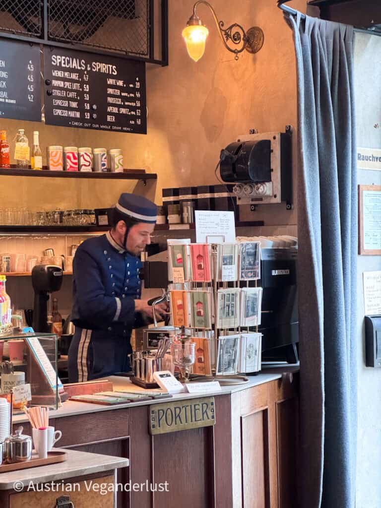 Barista dressed in a vintage porter-style uniform prepares drinks behind a café counter labeled “Portier,” surrounded by postcards, a specials board, and warm retro décor.