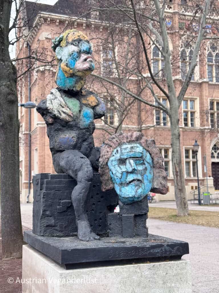 Eine abstrakte Skulptur mit markanter Struktur und Farbgebung, die eine sitzende Figur mit überzeichneten Gesichtszügen und einem abgesetzten blauen Kopf zeigt, der an Beethoven erinnert; sie steht auf einem Steinsockel vor einem historischen Backsteingebäude.