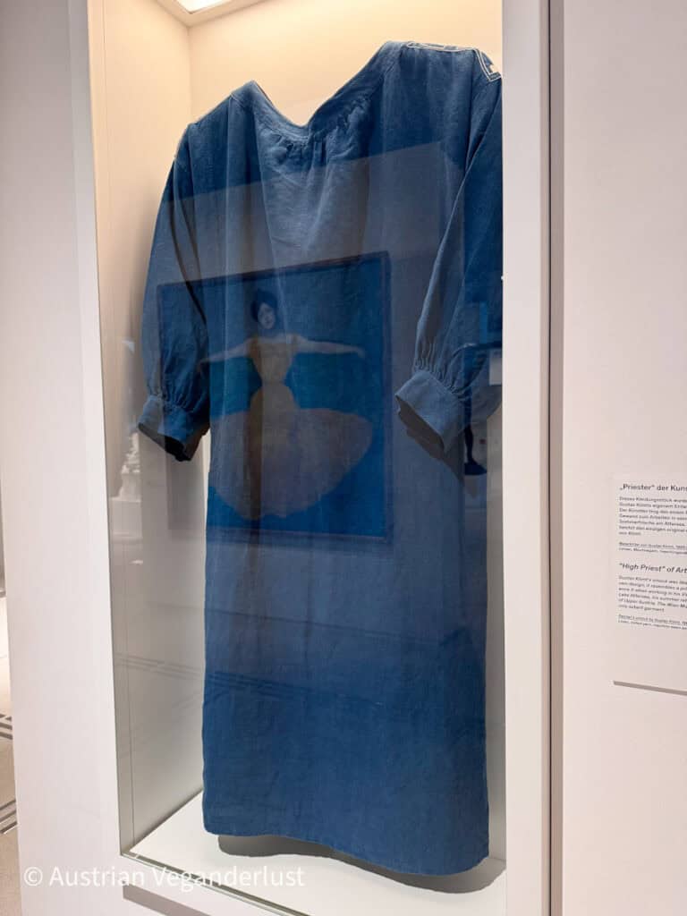 In einer Glasvitrine im Museum ist ein locker sitzender blauer Malerkittel mit weiten Ärmeln ausgestellt. Das schlichte Kleidungsstück reicht bis unter die Knie und wird von oben beleuchtet. Ein Wandschild neben der Vitrine verweist auf den „Priester“ der Kunst und weist darauf hin, dass der Kittel Gustav Klimt gehörte. Im Glas sind schwach die Spiegelungen der umliegenden Kunstwerke zu erkennen.