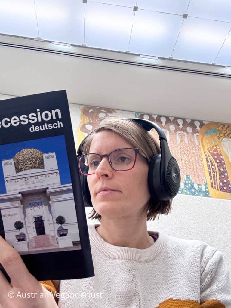 Eine Frau mit Kopfhörern liest einen deutschsprachigen Reiseführer mit dem Titel „Secession deutsch“, auf dessen Cover das Secessionsgebäude zu sehen ist, während sie drinnen sitzt und hinter ihr ein Teil von Gustav Klimts Beethoven-Fries zu sehen ist.