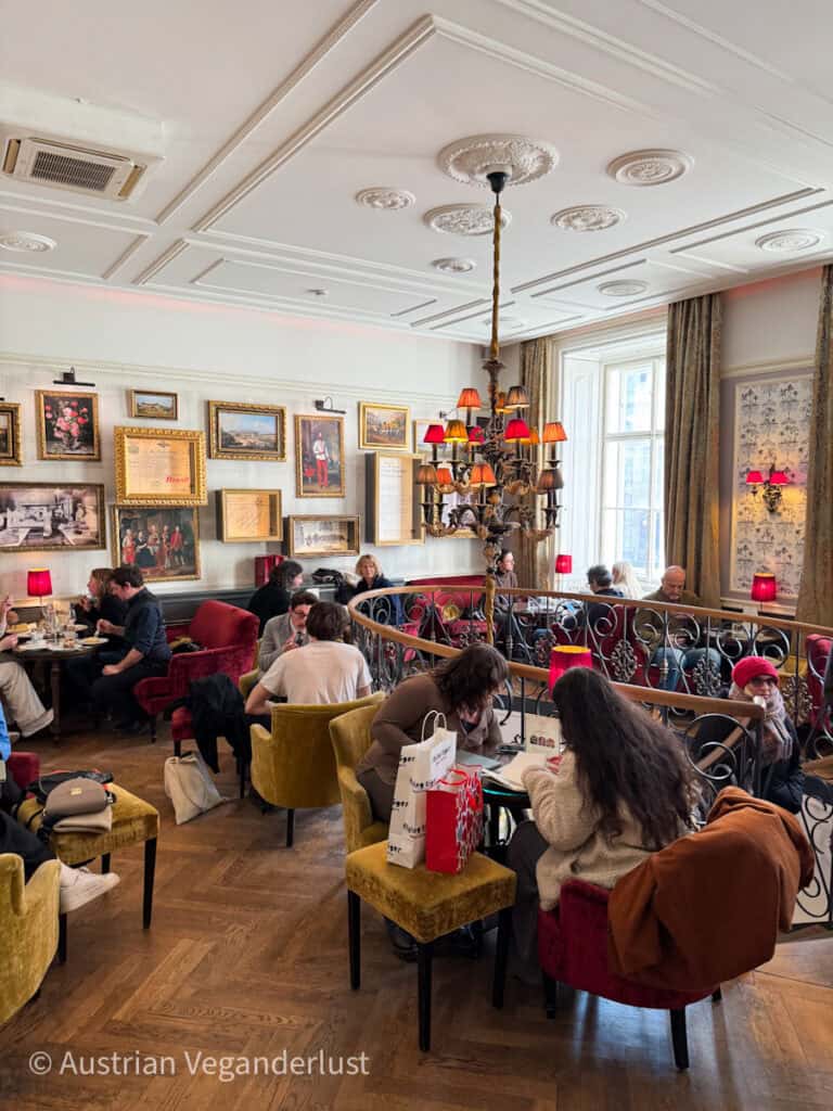 Lively café filled with guests seated in velvet chairs beneath a mix of traditional and quirky chandeliers, surrounded by framed vintage art and documents on white paneled walls.