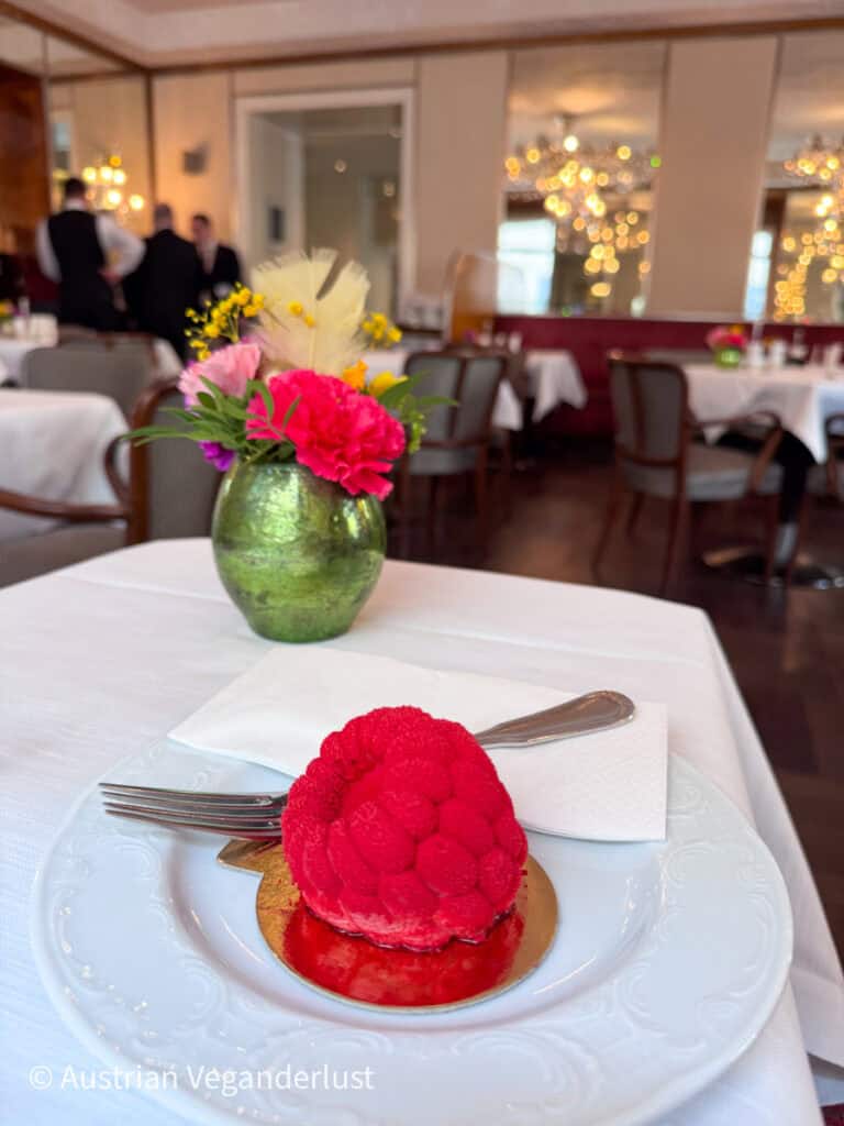 Kleines rundes Dessert mit frischen Himbeeren in leuchtendem Rot, angerichtet auf einem weißen Teller mit Gabel und Serviette, auf einem gedeckten Tisch in einem eleganten Café mit Blumenstrauß und Gästen im Hintergrund.