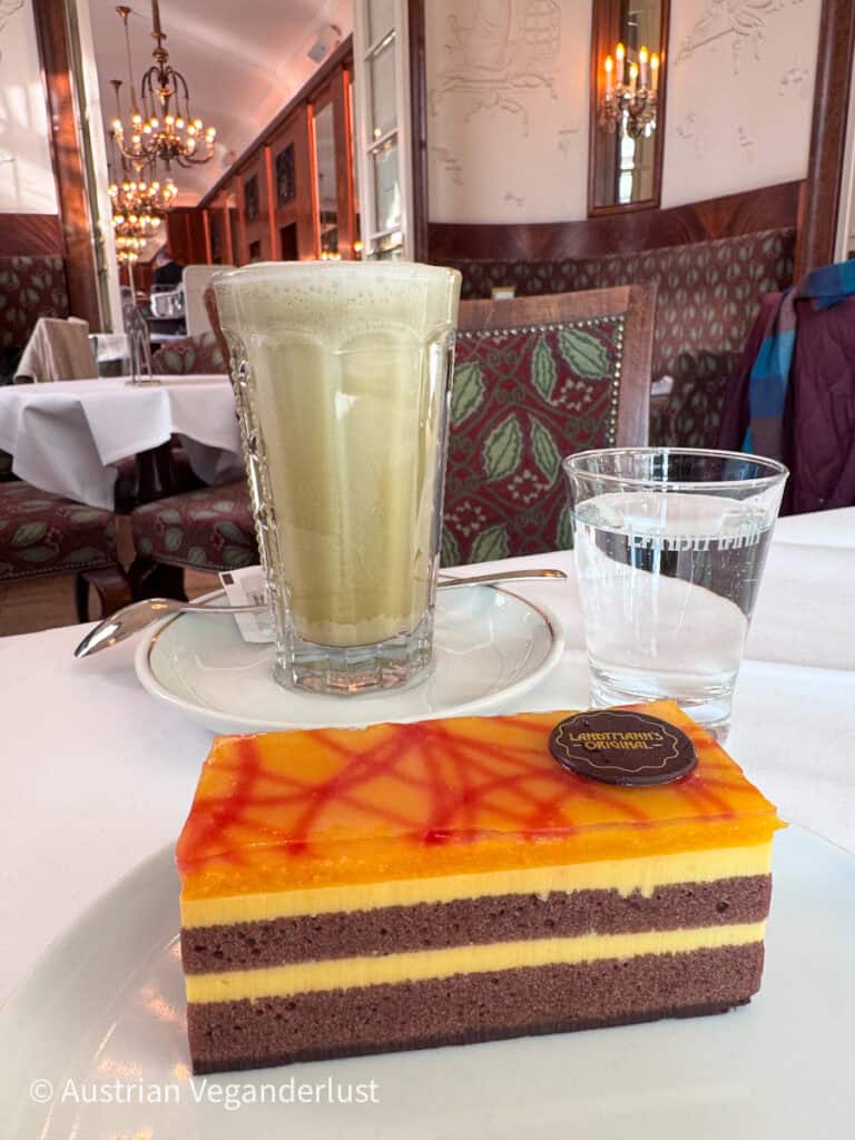 Stück Schichttorte mit gelber Creme und orangefarbener Glasur mit roten Linien, serviert auf einem Teller mit einem Glas Wasser und einem hohen Glas Kaffeegetränk in einem eleganten Wiener Kaffeehaus mit Kronleuchtern im Hintergrund.