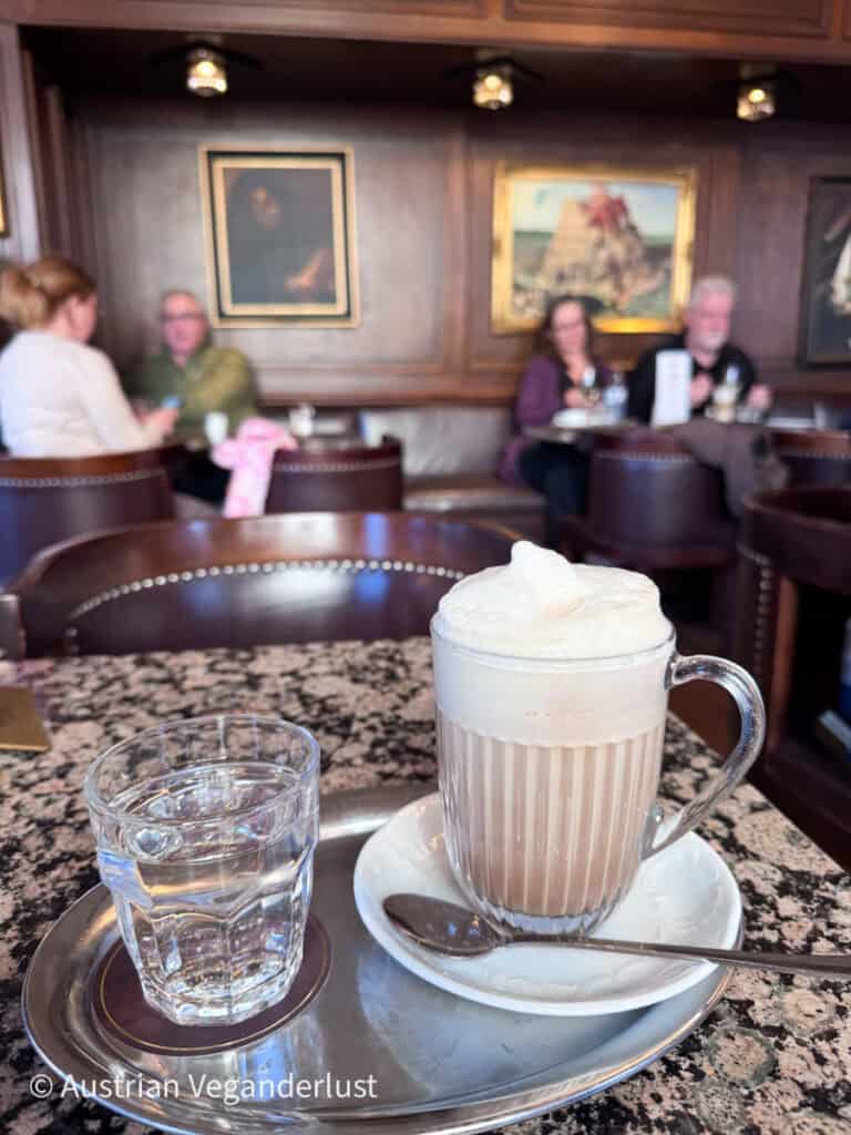 Glas Latte Macchiato mit dichtem Milchschaum auf einem Tablett neben einem Glas Wasser, serviert auf einem Marmortisch in einem klassischen Wiener Kaffeehaus mit Gästen im Hintergrund.