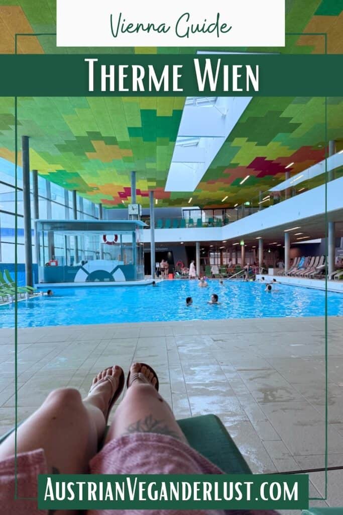 Indoor pool at Therme Wien with a colorful geometric ceiling, large windows, and people swimming, viewed from a lounge chair with legs stretched out in the foreground. Overlaid text reads "Vienna Guide Therme Wien" and "AustrianVeganwanderlust.com".