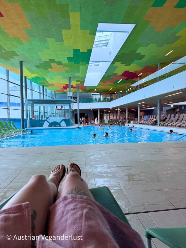 Spacious indoor pool with lounge chairs along the side and a colorful ceiling, seen from a reclined seating area inside a thermal bath.
