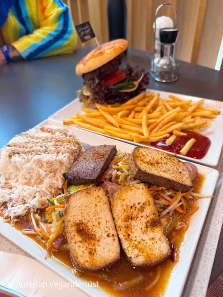 Meal with a veggie burger and fries alongside a plate of rice, stir fried vegetables, and tofu in sauce on a table.