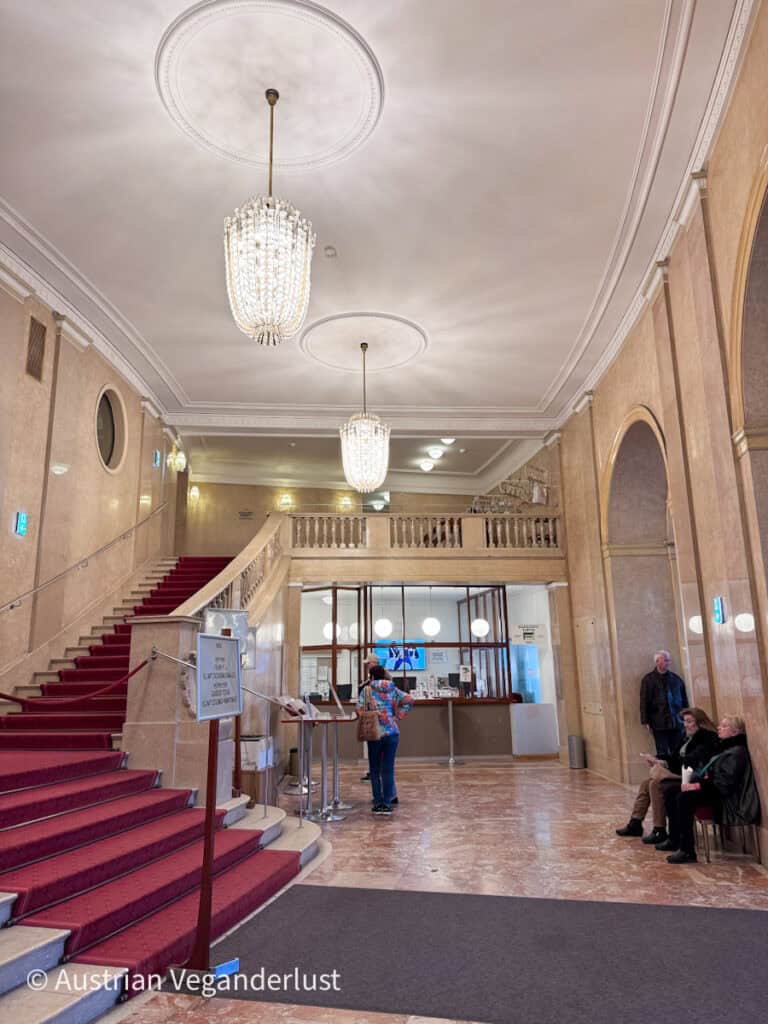 Die Lobby des Burgtheaters mit einer mit rotem Teppich ausgelegten Treppe, Kristallkronleuchtern und Besuchern, die in der Nähe eines Ticketschalters stehen. Auf einem Schild neben der Treppe steht „Kassa“ und „Information“.