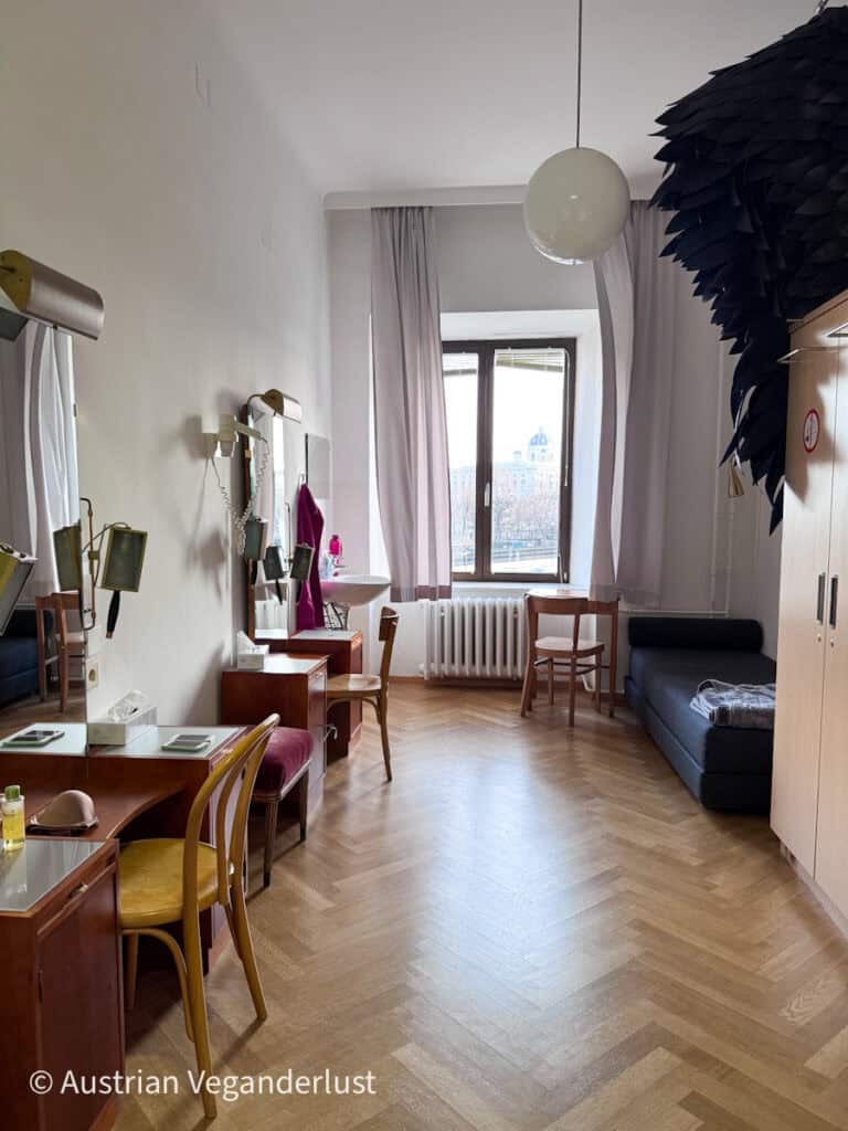 Ein schlichter Umkleideraum mit einem Holztisch, einem Spiegel, Stühlen und einem Fenster, durch das Tageslicht hereinfällt – ein ruhiger Ort hinter den Kulissen.