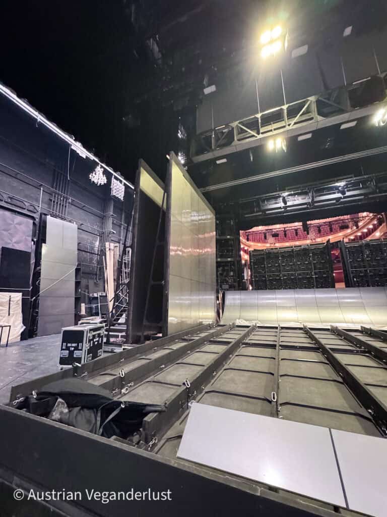 Ein Blick hinter die Kulissen einer Theaterbühne mit großen Kulissenelementen, Beleuchtungsanlagen und einem mechanischen Boden, der den technischen Aufbau offenbart.