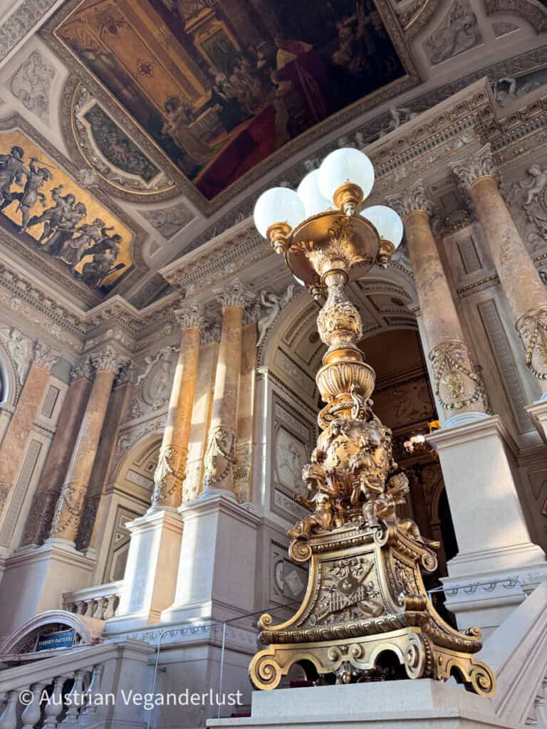 Ein kunstvoll gestalteter Innenraum mit imposanten Säulen, vergoldeten Skulpturen, bemalten Deckenpaneelen und einer aufwendig verzierten Lampe im Vordergrund.