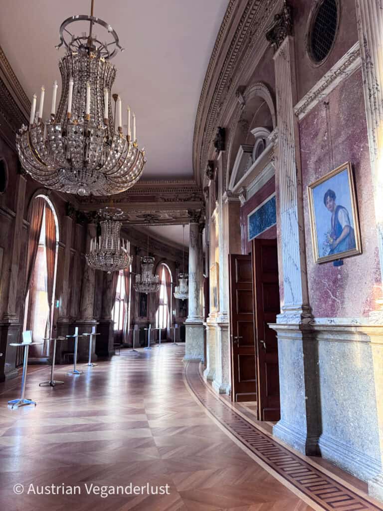 Ein prunkvoller Flur mit Kristallkronleuchtern, hohen Bogenfenstern, Marmorsäulen und gerahmten Porträts an den geschwungenen Wänden, der einen eleganten Treffpunkt vor der Vorstellung bildet.
