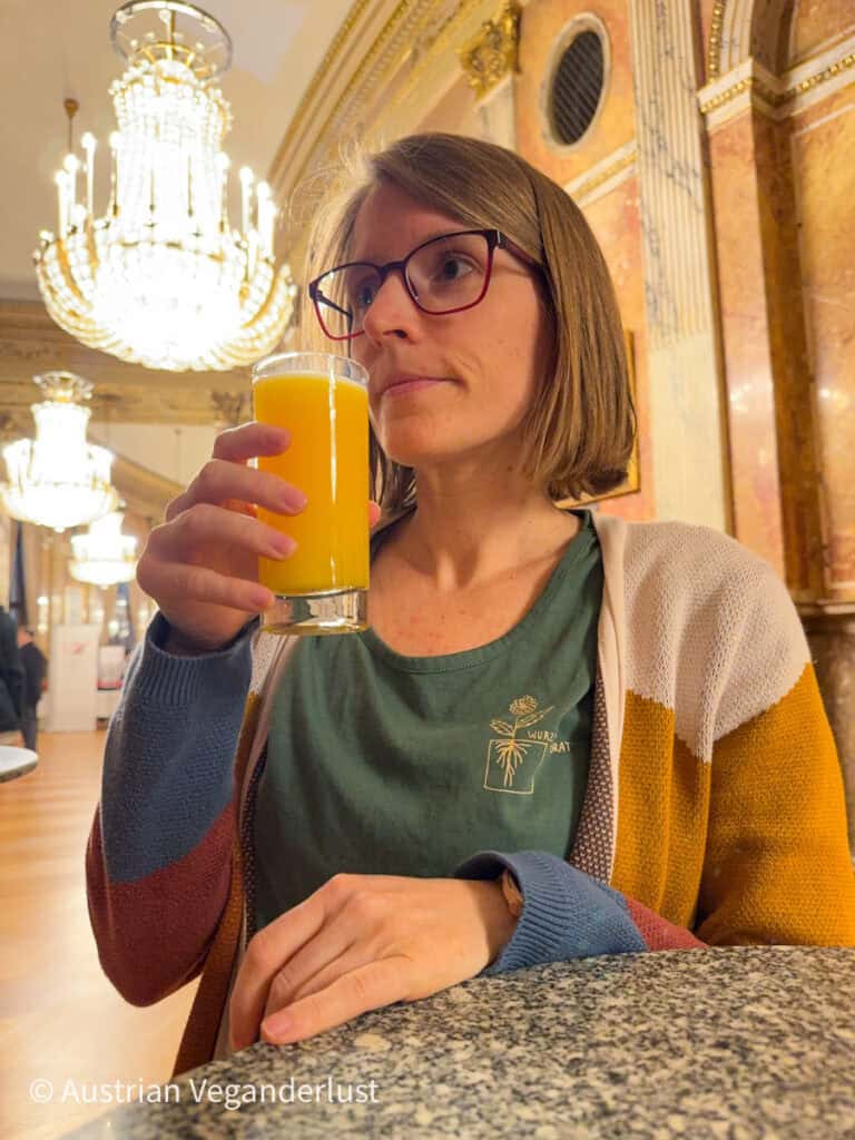 Tina hält ein Glas Orangensaft in der Hand, hinter ihr sind Kronleuchter und Marmorwände zu sehen – ein entspannter Moment bei einem Theaterbesuch.
