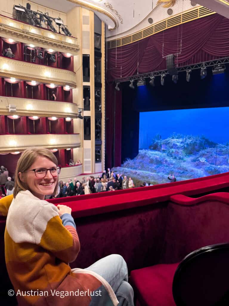 Tina lächelt von einem roten Samt-Balkonsitz im Burgtheater aus auf die Bühne mit ihrem blauen Bühnenbild, umgeben von kunstvoll verzierten Logenplätzen und warmer Theaterbeleuchtung.