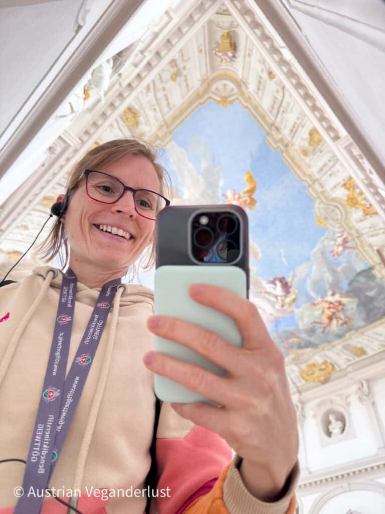 Tina wearing glasses taking a selfie while looking at their phone beneath an ornate ceiling with painted sky, clouds, and figures framed by decorative white and gold architecture.