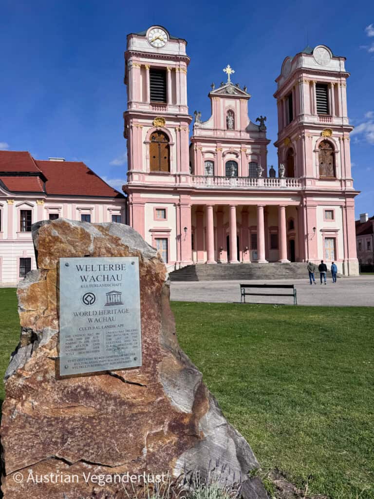 Pink baroque church facade with twin towers and a clock, seen from a grassy courtyard with a stone plaque reading "Welt­erbe Wachau Kulturlandschaft" and "World Heritage Wachau Cultural Landscape."