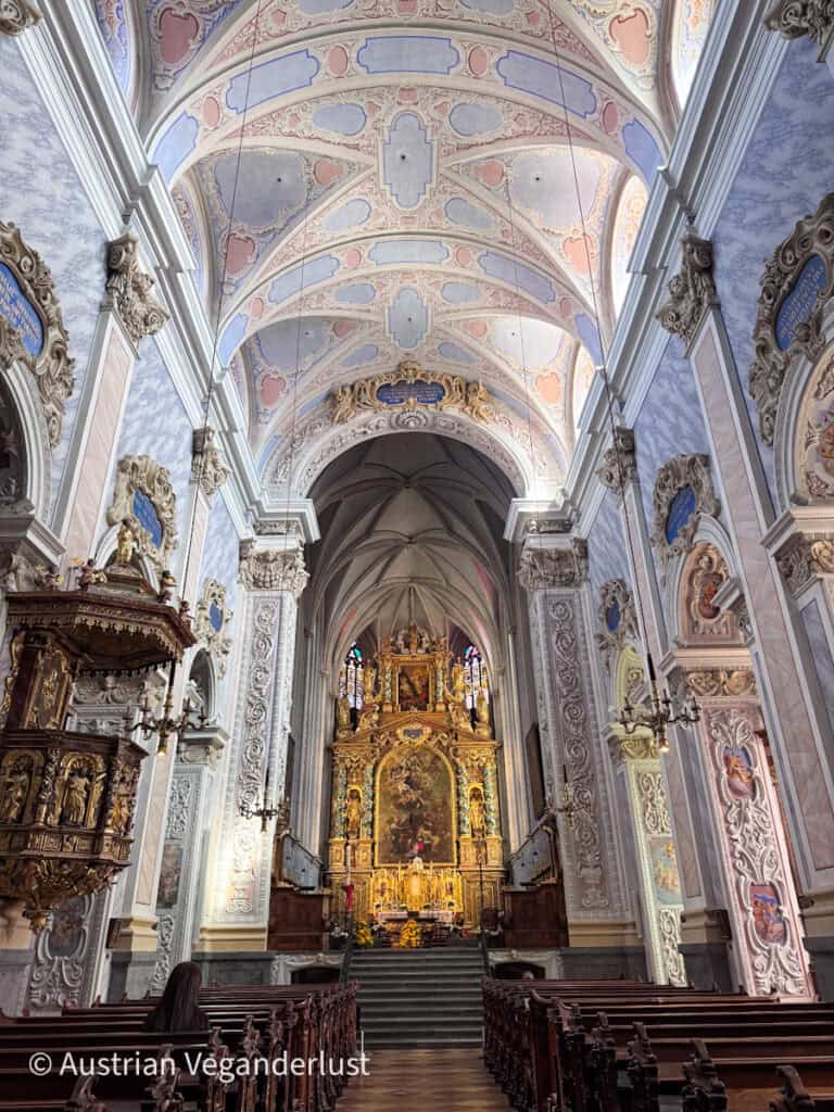 Interior of a grand church with high vaulted ceilings, pastel and gold detailing, and an elaborate altar at the far end framed by arches and rows of wooden pews.