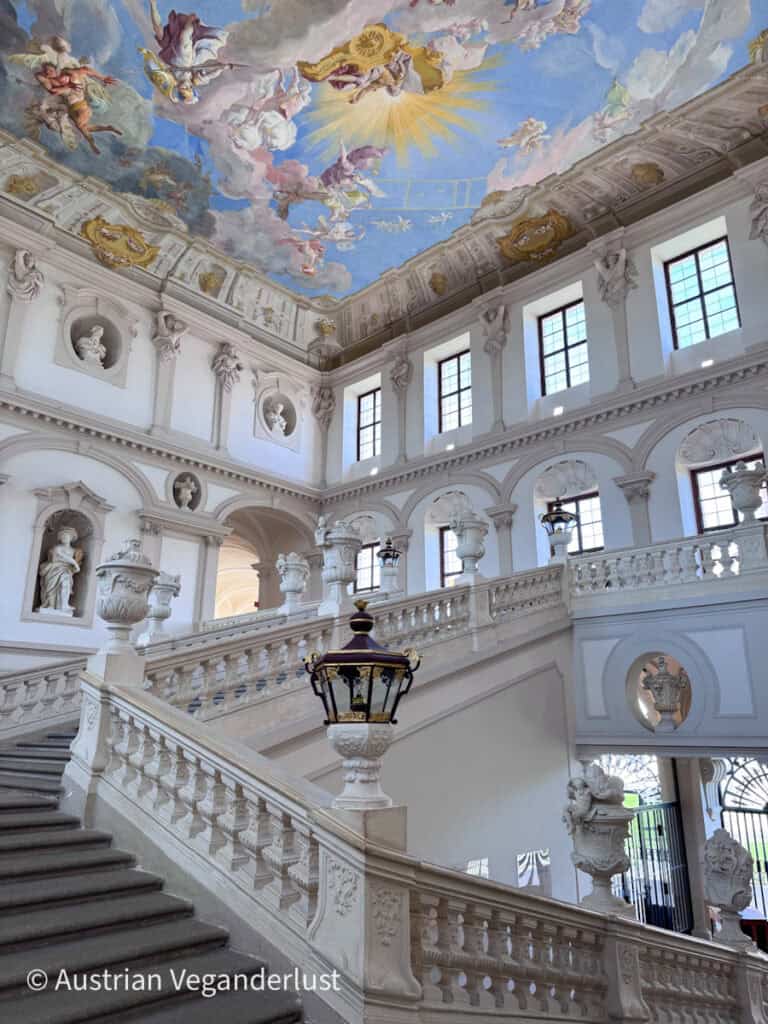 Grand interior staircase with white stone balustrades, statues, and large windows, leading up to a ceiling painted with colorful figures and golden details.