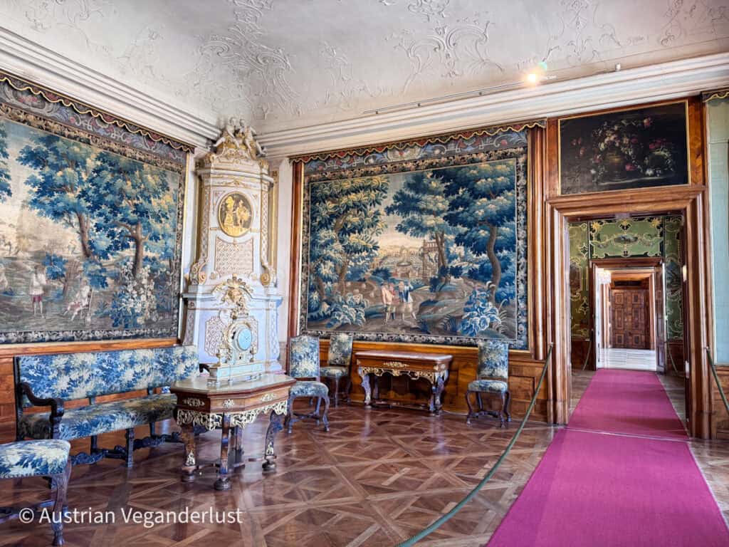 Elegant historic room with parquet flooring, upholstered chairs, ornate clock, and large tapestry wall hangings depicting pastoral scenes, with a red carpet leading through a doorway.
