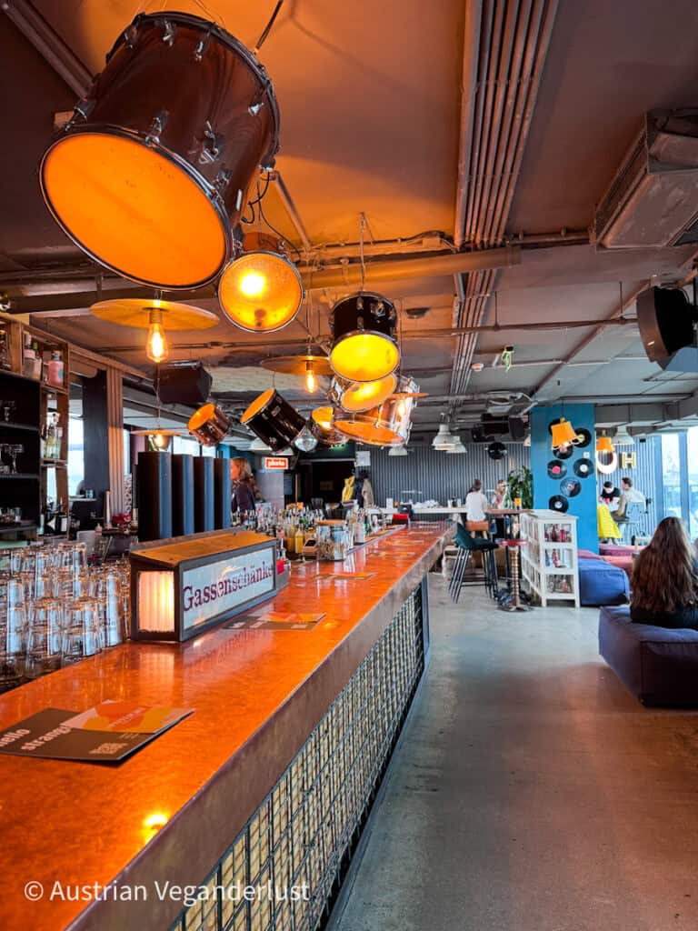 Trendiges Bar-Interieur mit einer langen Kupfertheke und hängenden Trommelleuchten, die in warmem Orange leuchten, während die Leute in einer entspannten Lounge-Atmosphäre sitzen und sich unterhalten.
