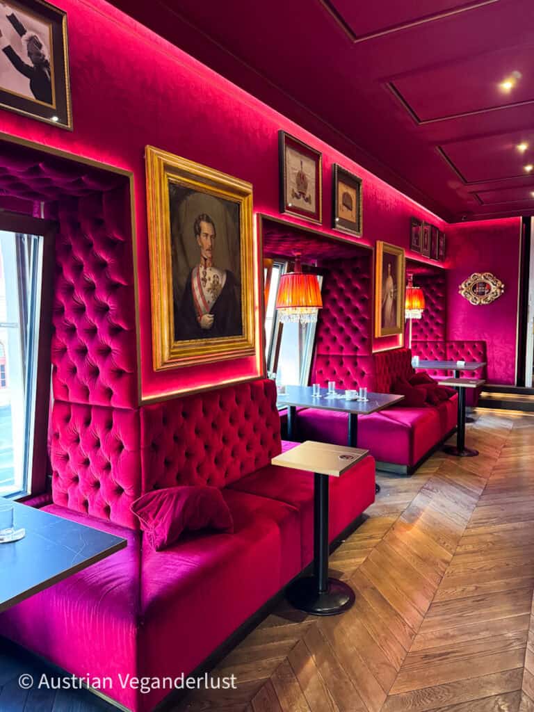 Interior of an elegant café with deep red velvet seating, framed portraits on the walls, small tables, and soft lighting creating a cozy atmosphere.