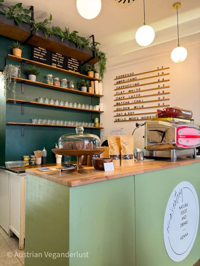 Minimalistischer Wiener Café-Tresen mit grünen Wänden, Holzregalen, Pflanzen und einer Speisekarte mit den Aufschriften „ESPRESSO 3,2 AMERICANO 3,9 CAPPUCCINO 4,4 FLAT WHITE 5,2 CAFÉ LATTE 4,9 MATCHA CHAI 5,2 GOLDEN MILK 5,2 HAFER-SOJA“ – daneben eine Kaffeemaschine und Kuchen unter einer Glasglocke.