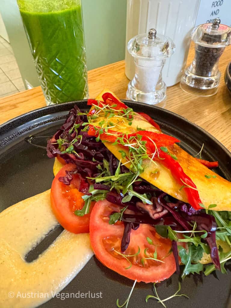 Ein veganes Omelett, gefüllt mit Tomaten, Rotkohl, Blattgemüse und Sprossen, serviert mit einer cremigen Soße und einem grünen Smoothie in einem hohen Glas auf einem Cafétisch.