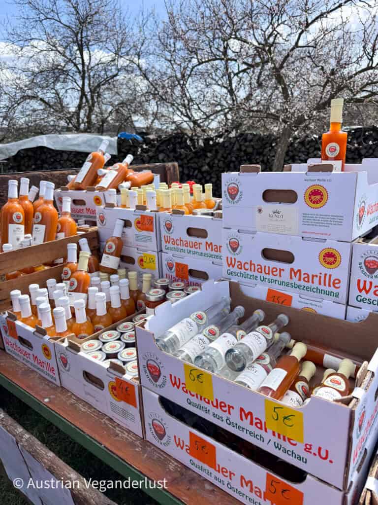 Outdoor market display of bottles and jars labeled "Wachauer Marille g.U." with price tags like "5€" and "4€" arranged in wooden crates.