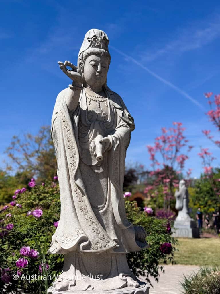 Stone statue of a robed figure standing among blooming pink flowers and greenery beneath a clear blue sky. The carved sculpture is framed by colorful garden plants and another statue in the distance.