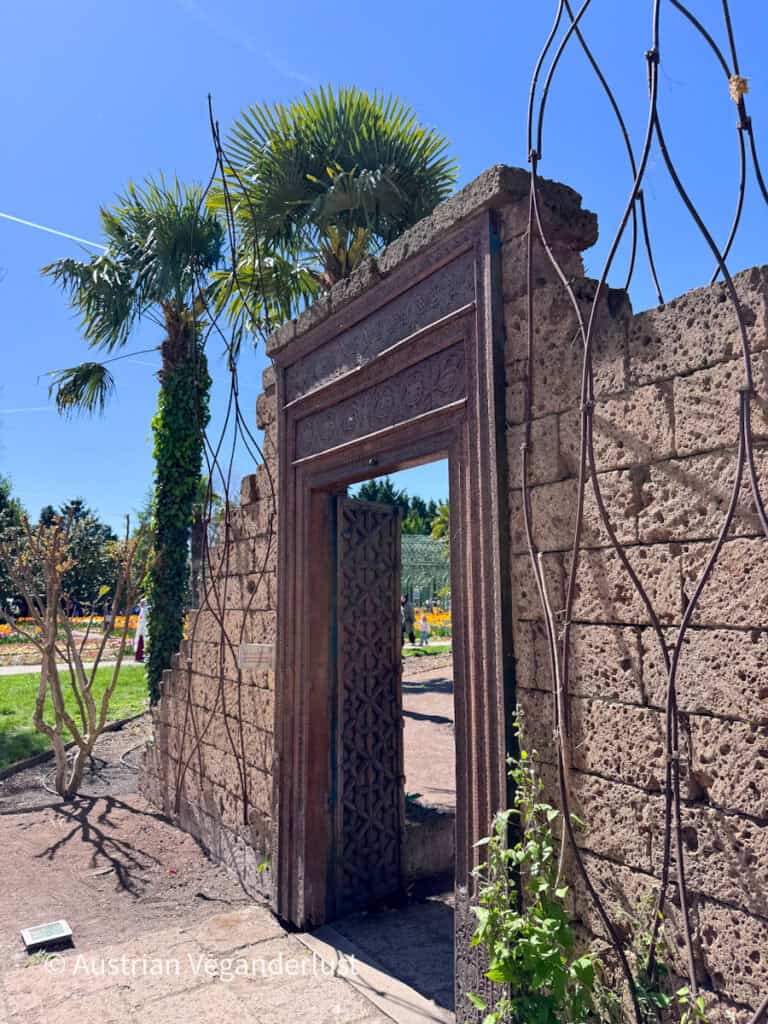 Weathered stone garden wall with an ornate carved doorway standing beneath tall palm trees and a bright blue sky. The open wooden door leads toward colorful flower beds and landscaped paths beyond.