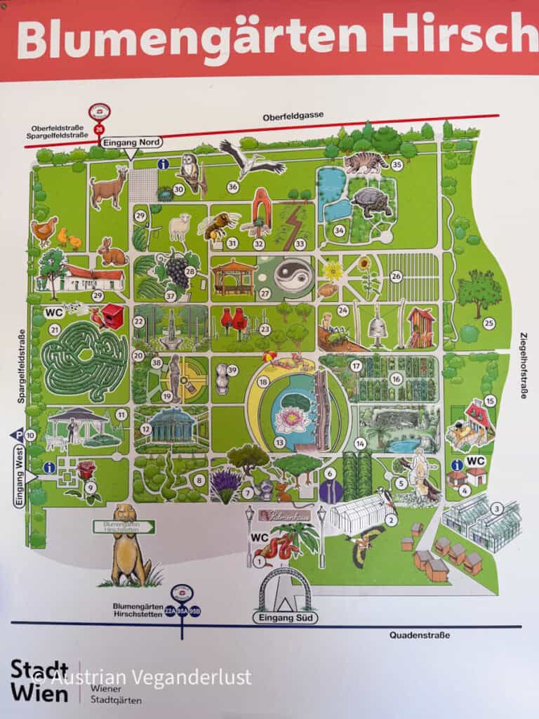 Illustrated map sign for "Blumengärten Hirschstetten" showing garden sections, pathways, animal illustrations, entrances, and visitor facilities. Text at the bottom reads "Stadt Wien Wiener Stadtgärten."