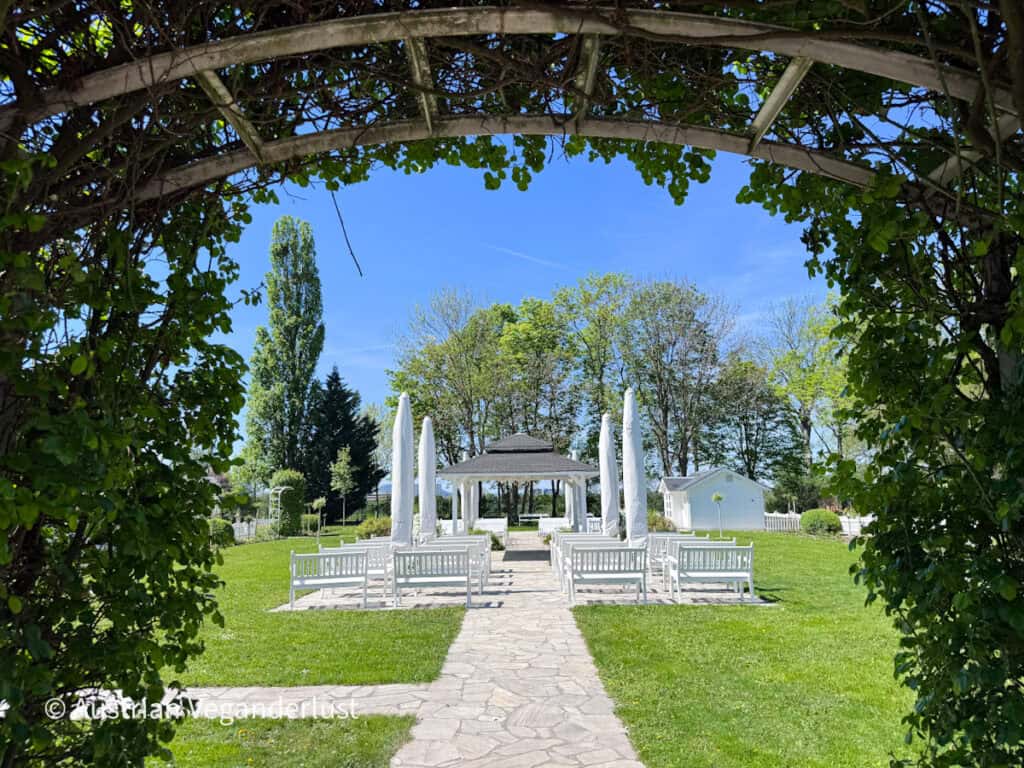 White garden gazebo and benches viewed through a leafy archway covered in green vines. A stone pathway leads across a neatly trimmed lawn toward the open pavilion under a clear sky.