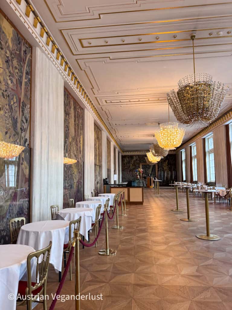 Long reception hall with parquet flooring, crystal chandeliers, and large tapestry covered walls beside neatly arranged dining tables. Warm lighting highlights the gold accents and spacious interior.