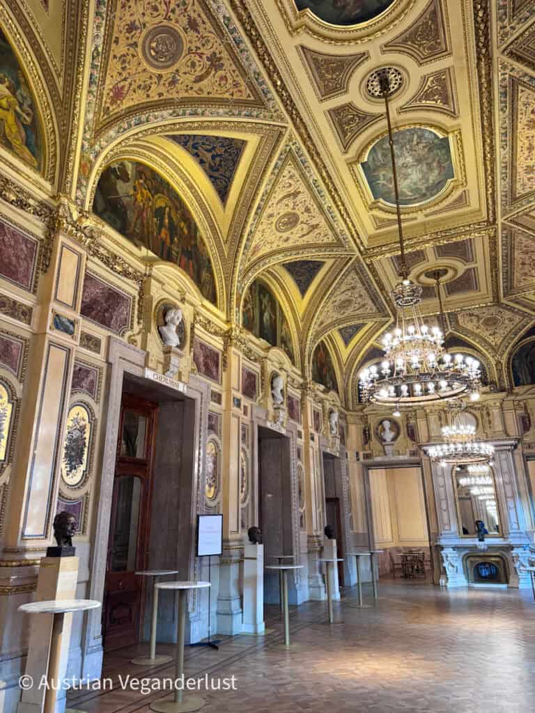 Lavishly decorated theater salon with painted arches, chandeliers, marble walls, and detailed gold trim across the vaulted ceiling. Bust sculptures line the walls beside tall mirrors and decorative fireplaces.