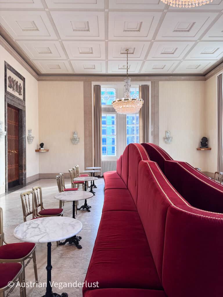 Elegant lounge area with tall red velvet seating, marble café tables, crystal chandeliers, and large windows letting in natural light. The pale walls and geometric ceiling create a bright and refined atmosphere.