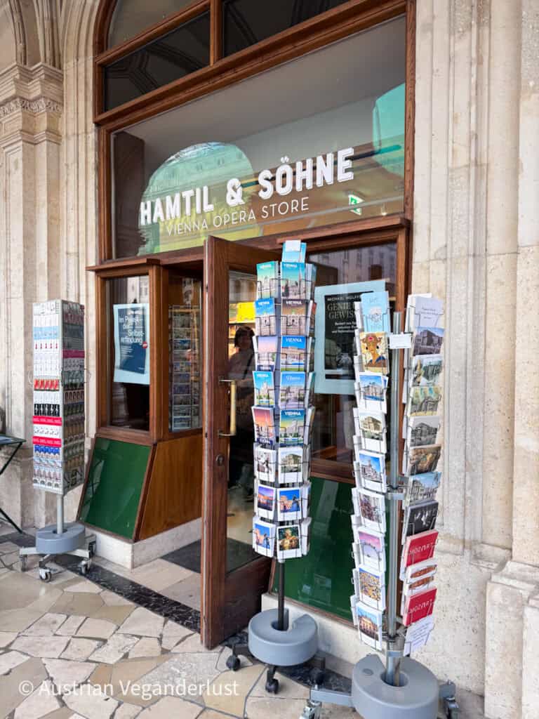 Entrance to the "HAMTIL & SÖHNE VIENNA OPERA STORE" with postcard racks and souvenir displays outside the wooden doorway. Brochures featuring Vienna landmarks line the storefront beneath arched stone architecture.