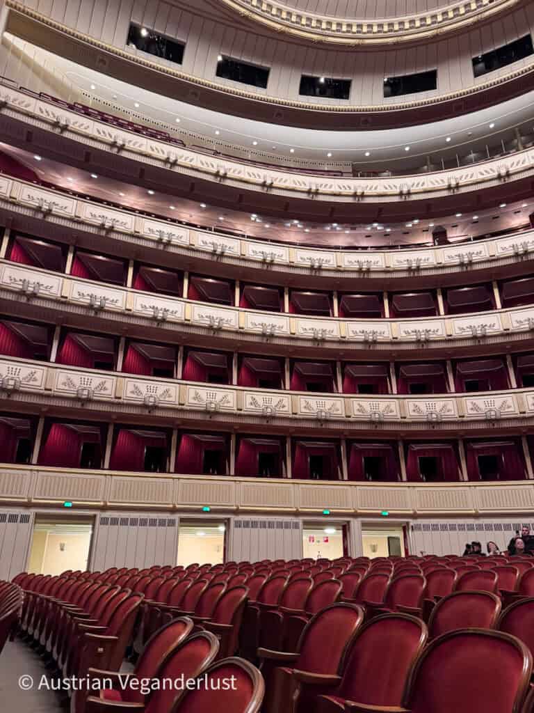 Empty opera house auditorium with rows of red velvet seats and several tiers of cream colored balconies with red curtains. The curved ceiling and ornate detailing emphasize the grand scale of the theater interior.