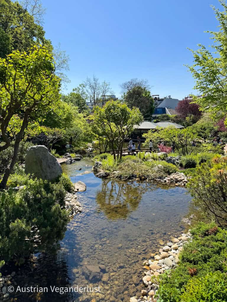 Ein klarer Bach schlängelt sich an einem sonnigen Tag durch einen japanischen Garten voller Felsen, Sträucher und Bäume. Im Hintergrund spazieren Besucher über Wege und über eine kleine Brücke.
