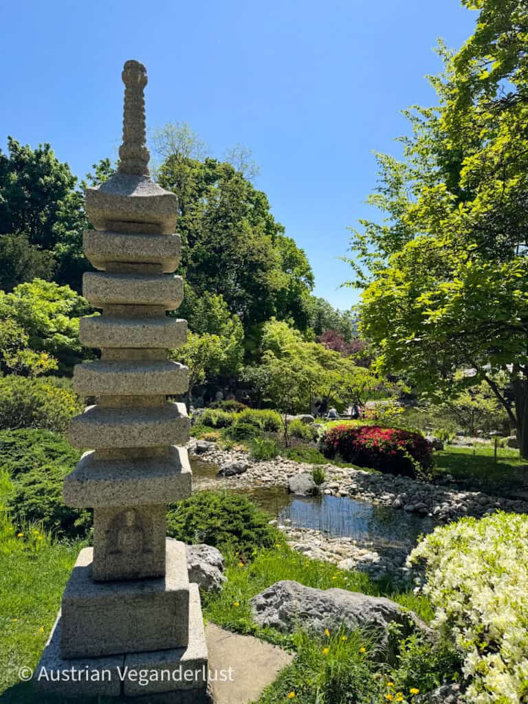 Ein hohes Pagoden-Denkmal aus Stein steht neben einem schmalen Bach in einem japanischen Garten. Blühende Sträucher, glatte Felsen und grüne Bäume umgeben das Wasser unter einem strahlend blauen Himmel.