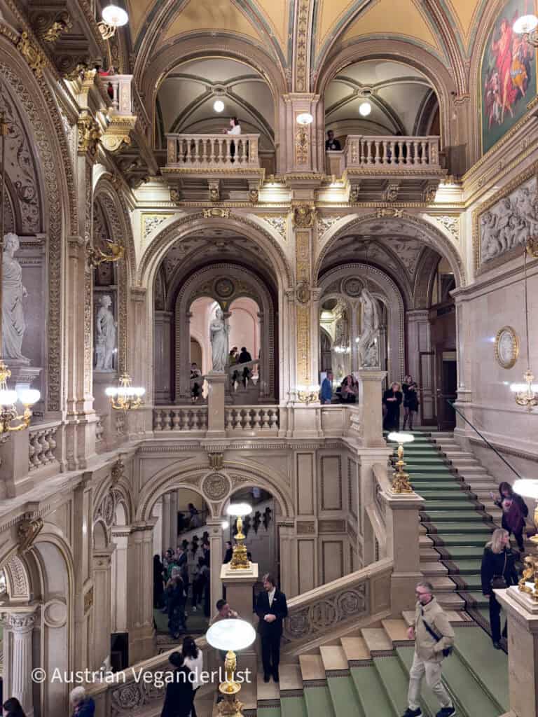Grand marble staircase inside an ornate opera house with vaulted ceilings, gold detailing, statues, and glowing chandeliers. Visitors walk along the green carpeted stairs and balconies beneath richly decorated arches and painted frescoes.