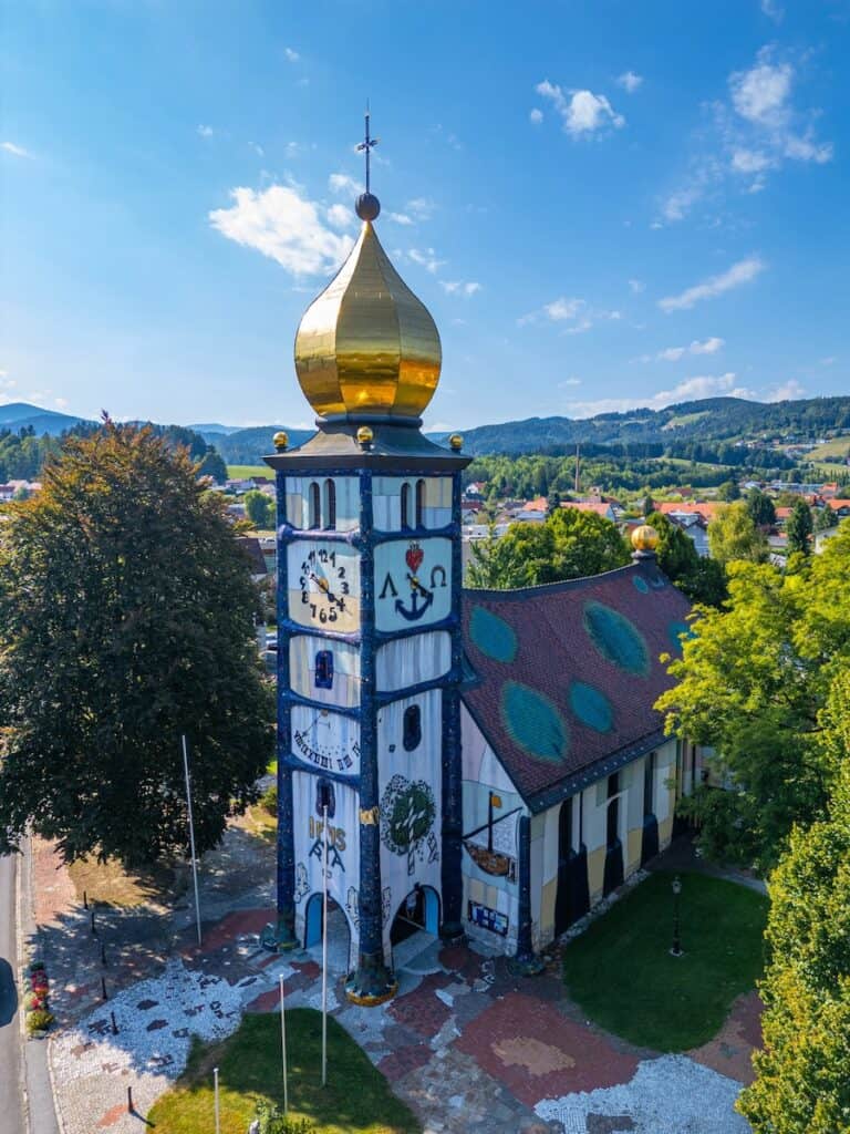 Church designed by Friedensreich Hundertwasser with a white facade, irregular decorative details, and a tall tower topped by a golden onion dome. The church stands among trees with rolling hills and a small town visible in the background on a sunny day.
