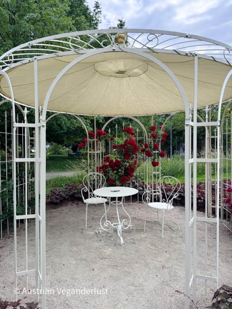 Ein weißer Pavillon aus Schmiedeeisen beherbergt einen kleinen runden Tisch und zwei dazu passende Stühle, umgeben von blühenden roten Rosen und Zierpflanzen. Die romantische Sitzecke befindet sich auf einer Kiesfläche unter einem dekorativen Baldachin in einem gepflegten Garten.