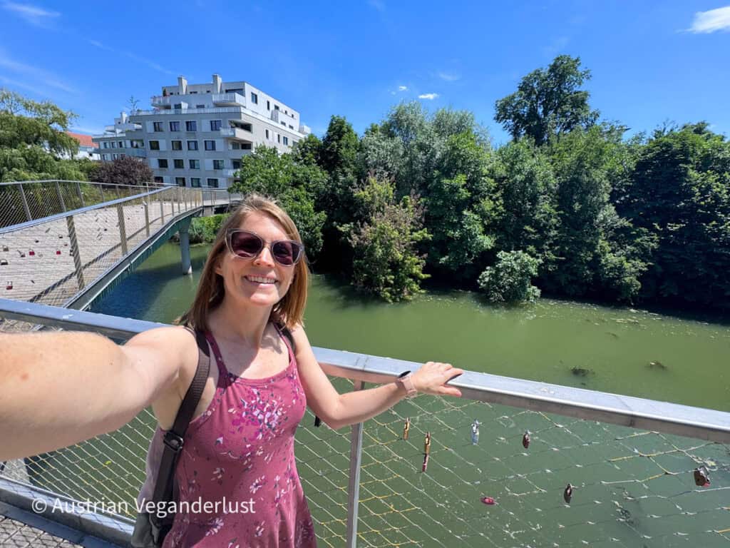 Eine lächelnde Frau mit Sonnenbrille steht an einem sonnigen Tag auf einer Fußgängerbrücke über einem grünen Fluss. Bäume säumen den Flusslauf, während moderne Wohnblocks und die geschwungene Brückenkonstruktion die Szene im Hintergrund einrahmen.