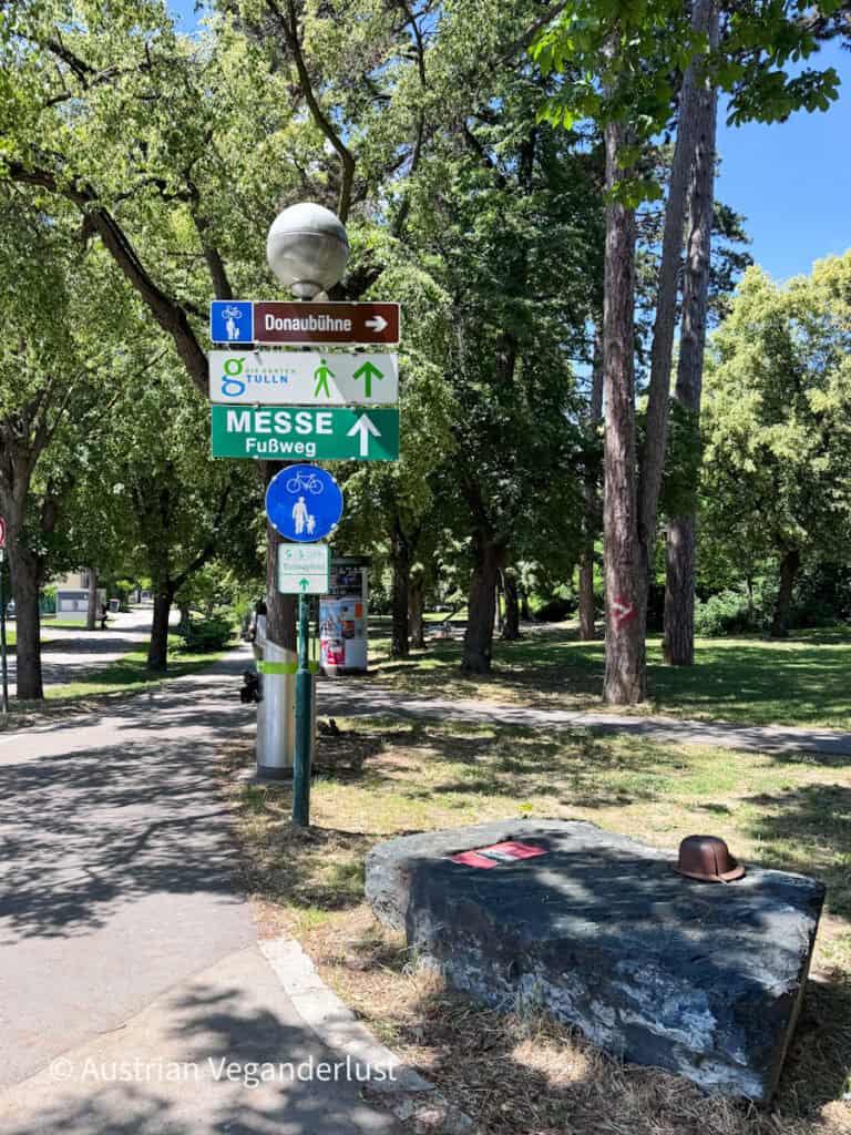 Wegweiser in einem schattigen Park am Flussufer weisen den Besuchern den Weg zur Donaubühne, zu „Die Garten Tulln“ und zum Fußgängerweg zur Messe. Hohe Bäume säumen die Fuß- und Radwege unter einem strahlenden Sommerhimmel.