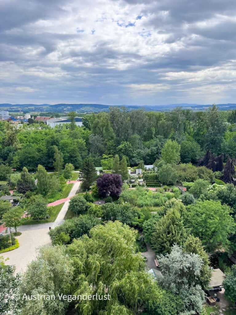 Weitreichender Blick aus der Vogelperspektive über „Die Garten Tulln“ mit gewundenen Wegen, Themengärten, altem Baumbestand und landschaftlich gestalteten Grünflächen, die sich bis zu den fernen Hügeln erstrecken. Über dem weitläufigen Gartenpark ziehen dunkle Wolken auf.