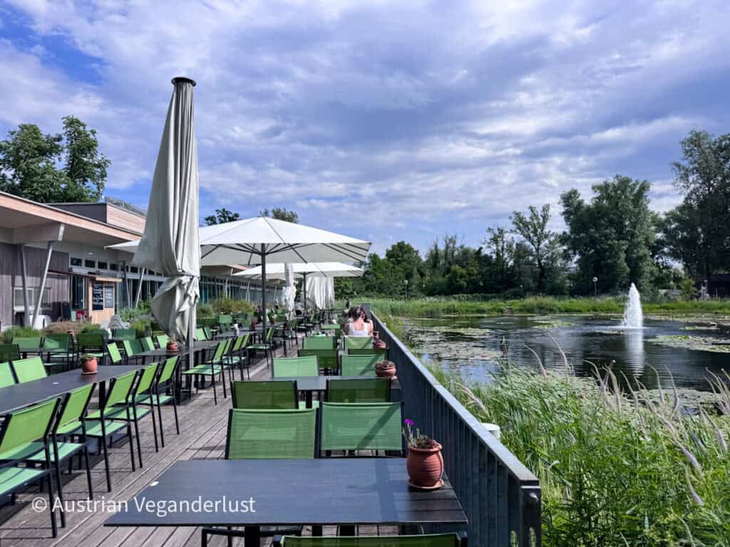 Die Terrasse des Restaurants im Freien mit grünen Stühlen und Tischen bietet einen Blick auf einen Teich voller Wasserpflanzen und einen Springbrunnen. An einem bewölkten Nachmittag spenden große Sonnenschirme am Wasser Schatten.